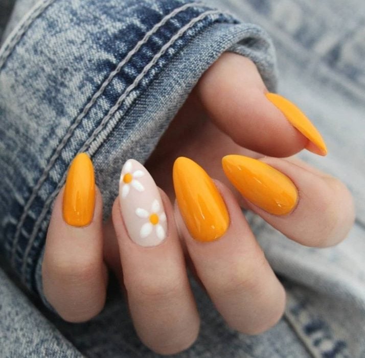 Mano de mujer con uñas largas en forma de almendra con una uñas pintadas con flores blancas, margaritas y las demás de color amarillo anaranjado para primavera