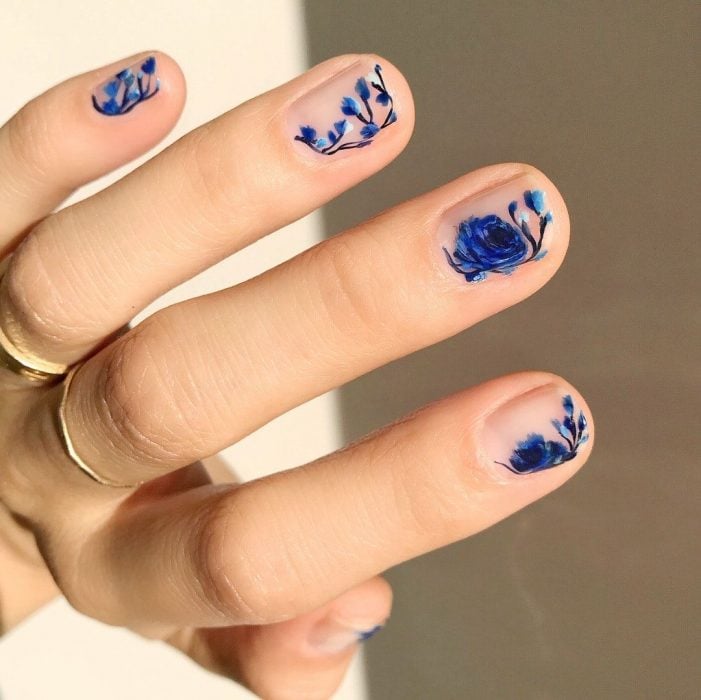 Mano de mujer con anillos dorados y uñas pintadas con flores azul rey sobre esmalte transparente