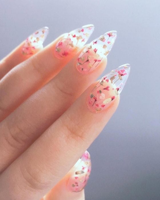 Mano de mujer con uñas largas en forma de almendra con esmalte transparente y pequeñas flores rosas y doradas