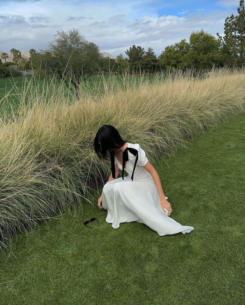 Kendall Jenner bending over to pick up sunglasses in field wearing white silk dress.