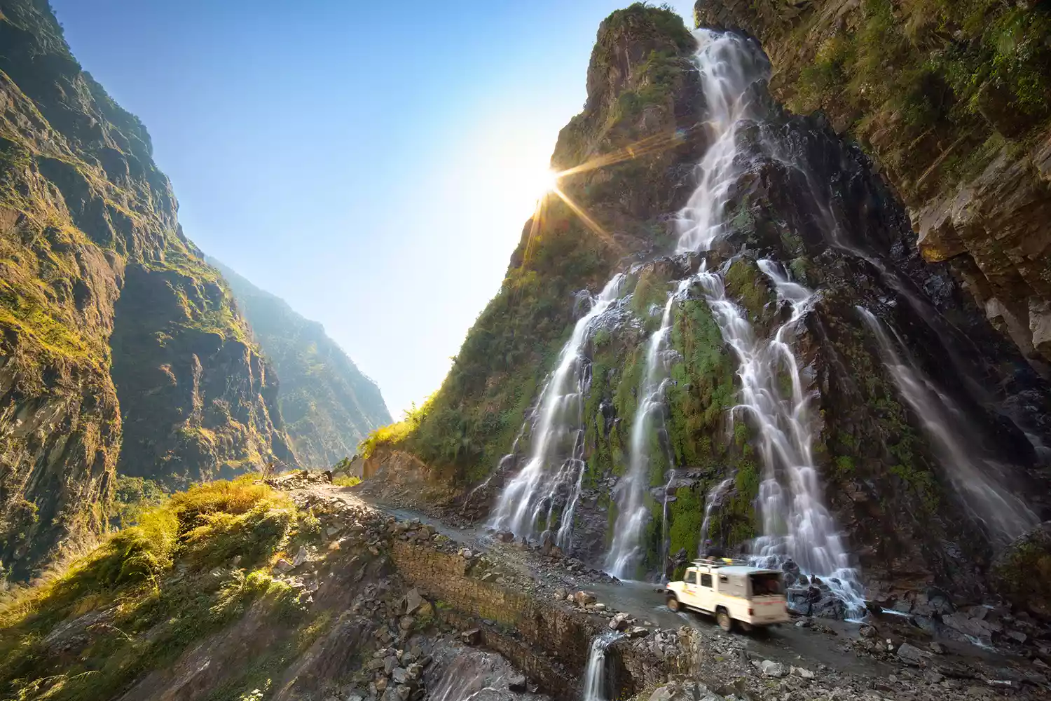 Car rides across the sunny mountain road beneath the waterfall in Nepal, Himalayas, Annapurna Conservation Area.